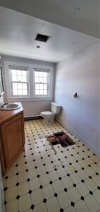 Partially renovated bathroom with shower removed, yellowed tiles on the floor, and light fixtures removed.